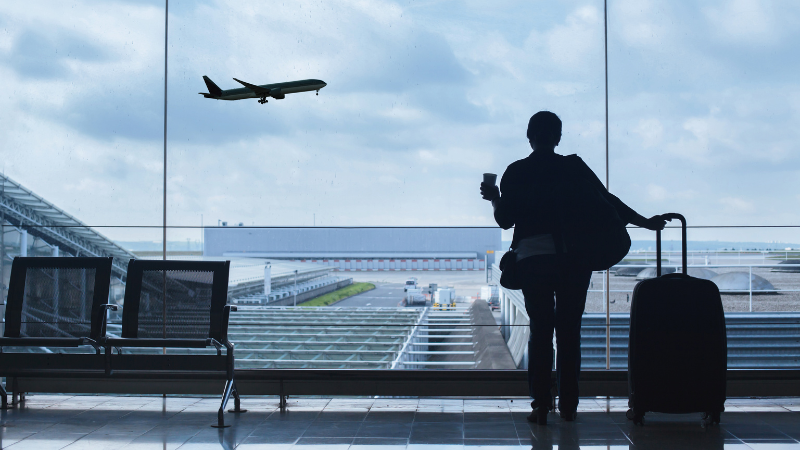 Femme de dos avec une valise, regardant les avions décoller à travers la vitre d’un aéroport